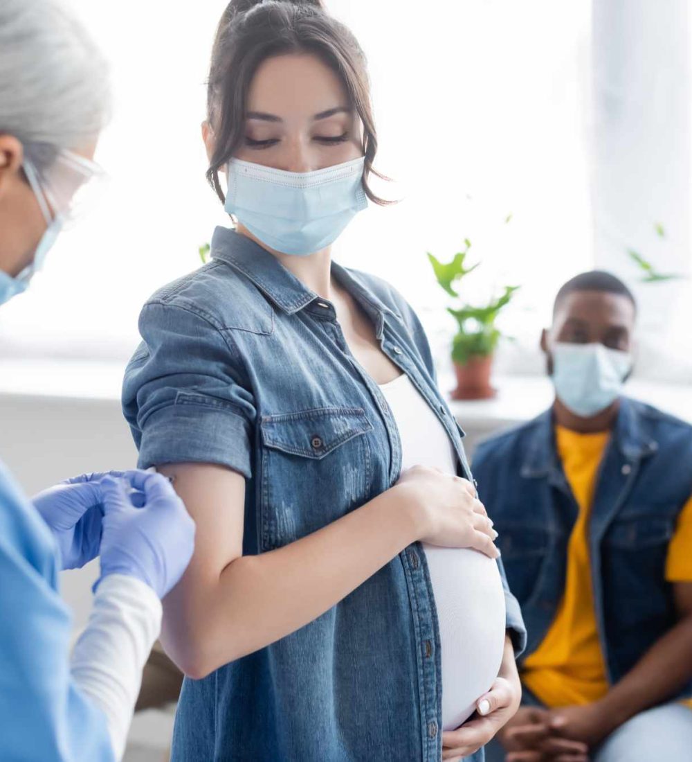 mature nurse vaccinating pregnant woman near blurred african american man in medical mask